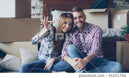 Portrait of happy married couple sitting together in new house, holding keys and looking at camera with carton boxes around. Family, relationship and relocation concept. Portrait of happy married couple sitting together in new house, holding keys and looking at camera with carton boxes around. Family, relationship and relocation concept. 95201740