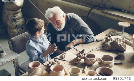 Curious little boy is kneading clay and talking to his grandfather professional potter while sitting together at table in workshop. Family tradition, professional pottery and childhood concept. Curious little boy is kneading clay and talking to his grandfather professional potter while sitting together at table in workshop. Family tradition, professional pottery and childhood concept. 95201748