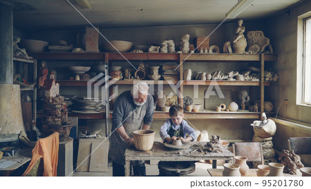 Skilled child is forming pot on potter's wheel while working with his retired grandfather in his figuline home studio. Professional equipment and beautiful ceramic things in background. 95201780