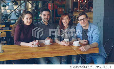Portrait of four smiling young people friends in casual clothes sitting at table in spacious cafe with tea cups and looking at camera. Eating out and friendship concept. 95201824
