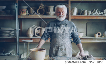 Portrait of senior silver-haired potter in muddy apron standing alone at table in workshop and looking at camera. Shelves with handmade vases and pots in background. Portrait of senior silver-haired potter in muddy apron standing alone at table in workshop and looking at camera. Shelves with handmade vases and pots in background. 95201855