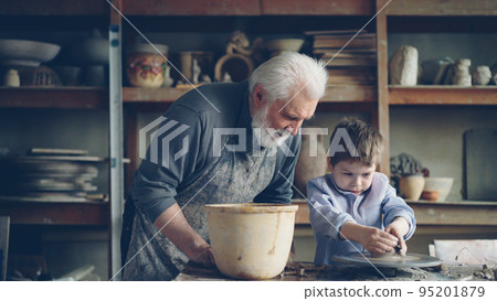 Pottery teacher senior adult in muddy apron is helping young student to form pot from piece of clay on throwing-wheel. Cute boy is awkward, and craftsman is helping him. 95201879