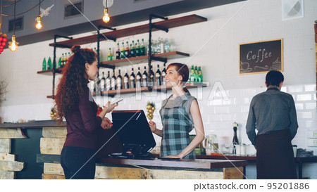Young people are buying coffee-to-go in nice local cafe and paying with smartphone while friendly workers are greeting customers, talking and selling drinks. 95201886