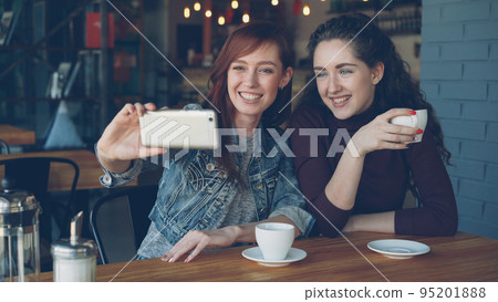 Pretty young girls best friends are taking selfie with smartphone then watching photos while drinking coffee in coffeehouse. Friendship, social media and having fun concept. Pretty young girls best friends are taking selfie with smartphone then watching photos while drinking coffee in coffeehouse. Friendship, social media and having fun concept. 95201888