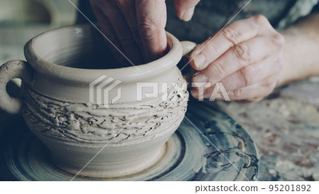 Close-up shot of muddy male potter's hands shaping and fixing ears of ceramic bowl on throwing wheel. Nimble craftsman is forming clay professionally creating beautiful clayware. 95201892