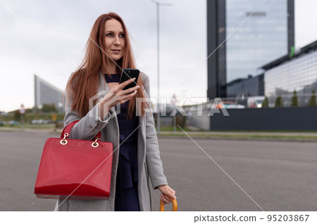 a red-haired businesswoman with a bright red bag stands with a mobile phone against the backdrop of a red-haired businesswoman with a bright red bag stands with a mobile phone against the backdrop of 95203867