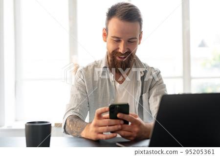 Handsome young bearded man spending time at home with phone in hands. Handsome young bearded man spending time at home with phone in hands. 95205361