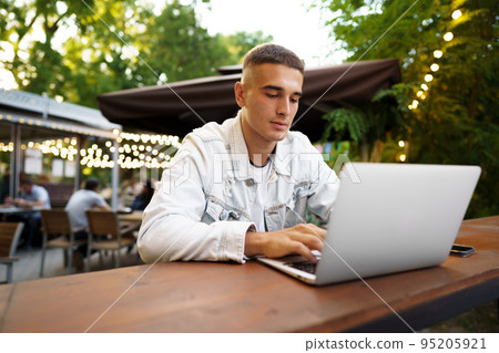 Young man sitting at table and typing on laptop keyboard while working in outdoor cafe 95205921
