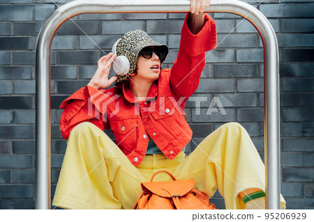 Hipster fashion young woman in bright clothes, sun glasses, bucket hat and ear phones sitting near gray brick wall background with bike stands frame. Urban city street fashion. Selective focus. 95206929