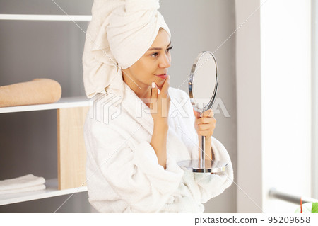 Woman with towel on head in bathroom looks and touches her face in the mirror and enjoys youth and hydration. Woman with towel on head in bathroom looks and touches her face in the mirror and enjoys youth and hydration. 95209658