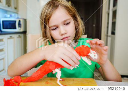 A blonde little girl makes a dinosaur craft out of wood and air plasticine.  95210940