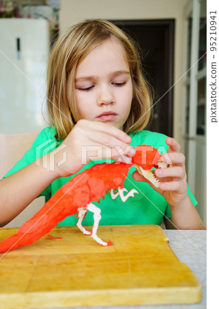 A blonde little girl makes a dinosaur craft out of wood and air plasticine.  95210941