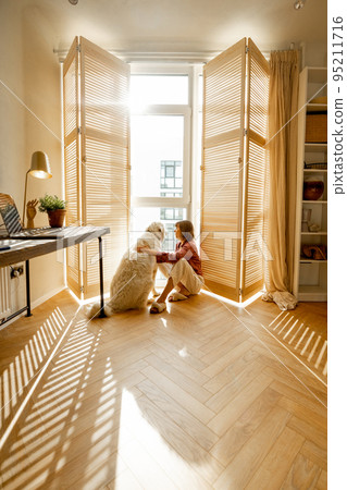 Woman sits with dog by the window blinds at home Woman sits with dog by the window blinds at home 95211716