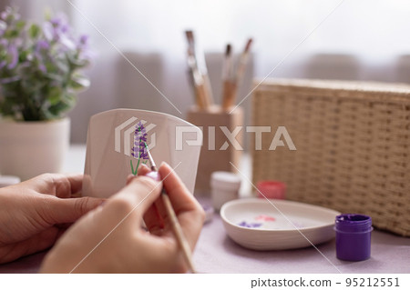 Female paints with a brush a sprig of a lavender flower, on a white ceramic pot 95212551
