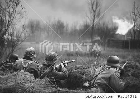 Re-enactors Armed Rifles And Dressed As World War Ii German Wehrmacht Infantry Soldiers Fighting Defensively In Trench. Defensive Position. Building On Fire On Background. Black And White Colors Re-enactors Armed Rifles And Dressed As World War Ii German Wehrmacht Infantry Soldiers Fighting Defensively In Trench. Defensive Position. Building On Fire On Background. Black And White Colors 95214081