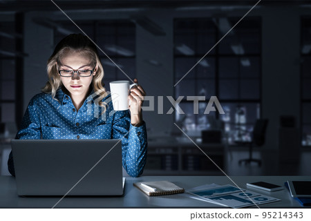 Attractive blonde working on laptop in dark office. Mixed media 95214343