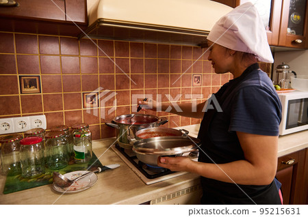 Rear view of a pretty woman, housewife in chef's apron and black apron, standing by a stove and cooking passata from organic tomatoes, making tomato sauce for preserving in sterilized cans for winter Rear view of a pretty woman, housewife in chef's apron and black apron, standing by a stove and cooking passata from organic tomatoes, making tomato sauce for preserving in sterilized cans for winter 95215631