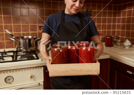 Selective focus on sterilized tins with freshly canned homemade organic tomato juice or passata, in a wooden crate, in the hands of a pretty woman housewife, standing against home kitchen background Selective focus on sterilized tins with freshly canned homemade organic tomato juice or passata, in a wooden crate, in the hands of a pretty woman housewife, standing against home kitchen background 95215635