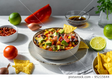 Cowboy caviar is traditional Mexican vegetable salad in bowl with ingredients and nachos on white wooden table with ingredients Cowboy caviar is traditional Mexican vegetable salad in bowl with ingredients and nachos on white wooden table with ingredients 95215644