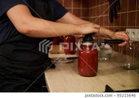 Close-up. Selective focus on a chef's hands using seamer or special seaming key, close lids of jars with freshly canned tomato sauce, juice or passata. Canning homegrown organic tomatoes for winter Close-up. Selective focus on a chef's hands using seamer or special seaming key, close lids of jars with freshly canned tomato sauce, juice or passata. Canning homegrown organic tomatoes for winter 95215646