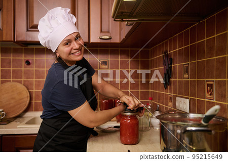 Charming housewife in chef's hat and apron, standing at a kitchen countertop, closing lids of jars with freshly brewed tomato juice, using a special seaming key, smiling cutely looking at the camera Charming housewife in chef's hat and apron, standing at a kitchen countertop, closing lids of jars with freshly brewed tomato juice, using a special seaming key, smiling cutely looking at the camera 95215649