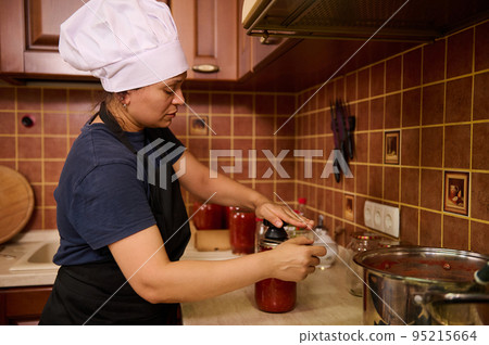 Side portrait of housewife closing lid on the jars with canned freshly boiled tomato juice or passata, with a seamer or special seaming key. Canning. Pickling. Cooking homemade tinned food. Marinating Side portrait of housewife closing lid on the jars with canned freshly boiled tomato juice or passata, with a seamer or special seaming key. Canning. Pickling. Cooking homemade tinned food. Marinating 95215664