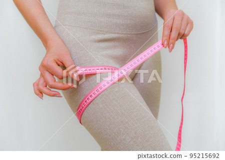Cropped view of slim woman measuring her leg with tape measure at home, close-up. An unrecognizable European woman checks the result of a weight loss diet or liposuction indoors. Healthy lifestyle. Cropped view of slim woman measuring her leg with tape measure at home, close-up. An unrecognizable European woman checks the result of a weight loss diet or liposuction indoors. Healthy lifestyle. 95215692