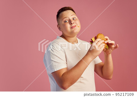 Half-length portrait of cute overweight boy in white t-shirt tasting delicious burger isolated on pink background. Fast food, taste, body positive, emotions 95217109