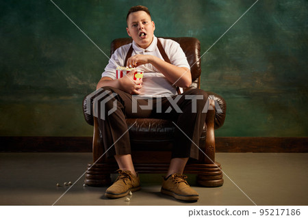 Portrait of fat teen boy wearing retro style clothes sitting in brown armchair on dark vintage background. Watching TV, eats popcorn. Overweight, carefree. 95217186