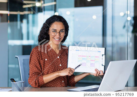Portrait Hispanic business coach, woman smiling and looking at camera, showing documents with economic indicators to camera, teacher teaching remotely, mentor using laptop for online learning. 95217769
