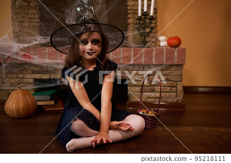 Cute little girl with Halloween themed make-up, dressed as a witch in a wizard's hat, looks at camera, sitting against the backdrop of a fireplace covered with cobwebs, pumpkins and old mystical books Cute little girl with Halloween themed make-up, dressed as a witch in a wizard's hat, looks at camera, sitting against the backdrop of a fireplace covered with cobwebs, pumpkins and old mystical books 95218111