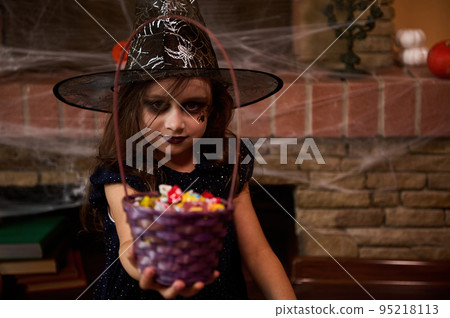 Cute little girl with Halloween themed make-up, dressed as a witch in a wizard's hat, looks at the camera and hands a basket of candies, sits on the background of a fireplace, covered with cobwebs. Cute little girl with Halloween themed make-up, dressed as a witch in a wizard's hat, looks at the camera and hands a basket of candies, sits on the background of a fireplace, covered with cobwebs. 95218113