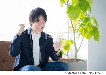 A man in his 20s looking at his smartphone and doing a guts pose-relaxing on the sofa in the living room 95218576