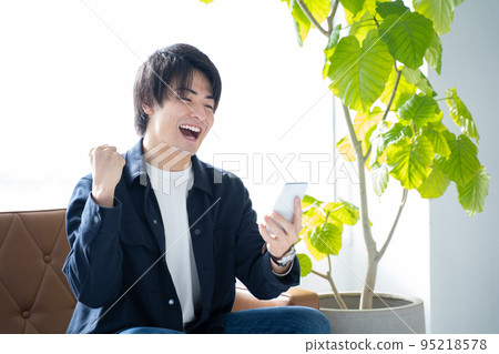 A man in his 20s looking at his smartphone and doing a guts pose-relaxing on the sofa in the living room A man in his 20s looking at his smartphone and doing a guts pose-relaxing on the sofa in the living room 95218578