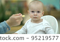 Mom feeds the baby puree from a small spoon. Mother and one year old son eating at the food court 95219577