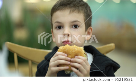 Tired sad boy of seven years old eats a burger at a food court after a school day. Child in sweatshirt enjoying a juicy burger Tired sad boy of seven years old eats a burger at a food court after a school day. Child in sweatshirt enjoying a juicy burger 95219581