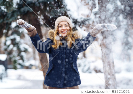 Beautiful girl playing with snow in winter forest. Smiling girl in a blue jacket and knitted hat. 95221061