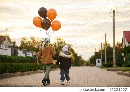 Rear view of elementary age kids, boy and girl or brother and sister, with orange black balloons, walking down the country street at sunset on Halloween. Autumn. Gothic festivity. October 31 95221967