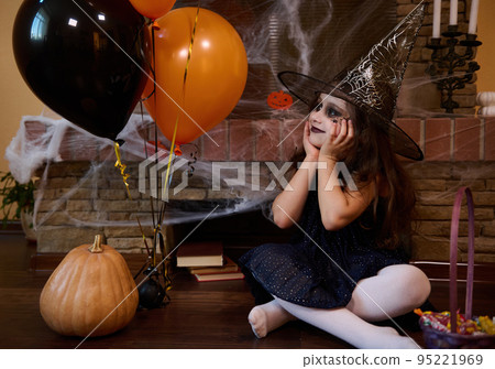 Beautiful little girl in a wizard's hat with face art makeup on her face, surrounded by black orange balloons, pumpkins and a cobweb-covered fireplace, enjoys Halloween party in a gothic home interior 95221969