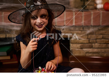 Adorable Caucasian little girl in witch carnival costume and wizard hat, with treat, smiling a beautiful smile sitting against a fireplace covered with spider web. Happy Halloween. Gothic festivity Adorable Caucasian little girl in witch carnival costume and wizard hat, with treat, smiling a beautiful smile sitting against a fireplace covered with spider web. Happy Halloween. Gothic festivity 95221993