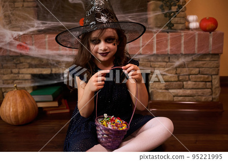 Little child girl dressed as a witch in black dress and wizard hat, sitting on the floor with a basket full of candies, against a fireplace covered with a spider web, pumpkin and old books. Halloween 95221995