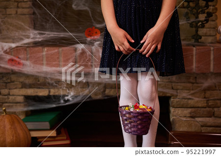 Cropped view. Selective focus. Legs of ittle child girl in black dress, with a basket full of candies, against a fireplace covered with spider web, pumpkin and old books. Halloween decoration at home Cropped view. Selective focus. Legs of ittle child girl in black dress, with a basket full of candies, against a fireplace covered with spider web, pumpkin and old books. Halloween decoration at home 95221997