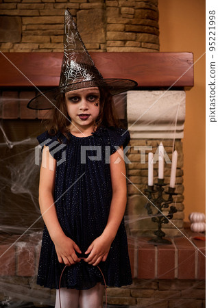 Charming little girl, a child witch in wizard hat, with a facial art makeup posing against a fireplace with spider web and candles on a candlestick. 31 October. Child celebrating Halloween. Autumn 95221998