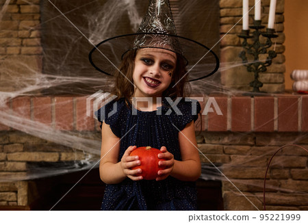 Funny child, Caucasian little girl with face art, wearing a wizard hat and black dress, smiling a beautiful toothy smile looking at camera, posing with a pumpkin against a fireplace with spider web Funny child, Caucasian little girl with face art, wearing a wizard hat and black dress, smiling a beautiful toothy smile looking at camera, posing with a pumpkin against a fireplace with spider web 95221999
