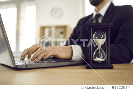 Man trying to meet deadline and working on laptop computer at desk with hour glass Man trying to meet deadline and working on laptop computer at desk with hour glass 95222674