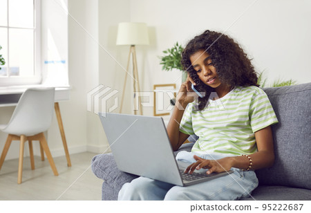 African america teen girl is calling mobile phone and using laptop sitting on couch at home. 95222687