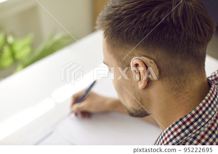 Hearing impaired student wearing modern hearing aid is sitting at desk and writing Hearing impaired student wearing modern hearing aid is sitting at desk and writing 95222695