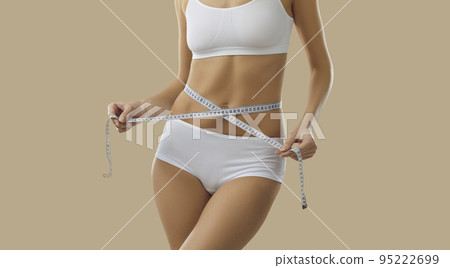 Slender woman in lingerie measures figure with tape measure standing on beige background. 95222699
