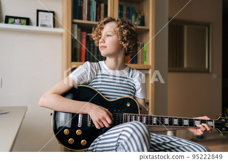 Portrait of pretty curly preteen girl sitting on chair and playing on electric guitar, learning new song at home, looking away. Cute little kid teaching to play guitar in bedroom with cozy interior. Portrait of pretty curly preteen girl sitting on chair and playing on electric guitar, learning new song at home, looking away. Cute little kid teaching to play guitar in bedroom with cozy interior. 95222849