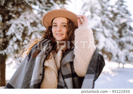 Smiling woman enjoying winter moments in snowy park. Young woman wearing hat, plaid scarf and coat. 95222850